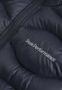 Námornícka prešívaná pufovaná bunda s lesklým nylónovým povrchom. Obsahuje zips a vyšívané logo "PeakPerformance" v šedej farbe.