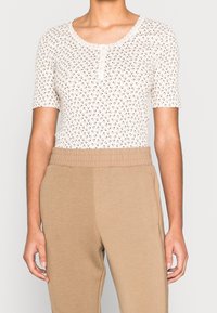 Persona que lleva una camisa blanca de manga corta con estampado floral y botones, junto con pantalones beige de cintura alta y cintura elástica.