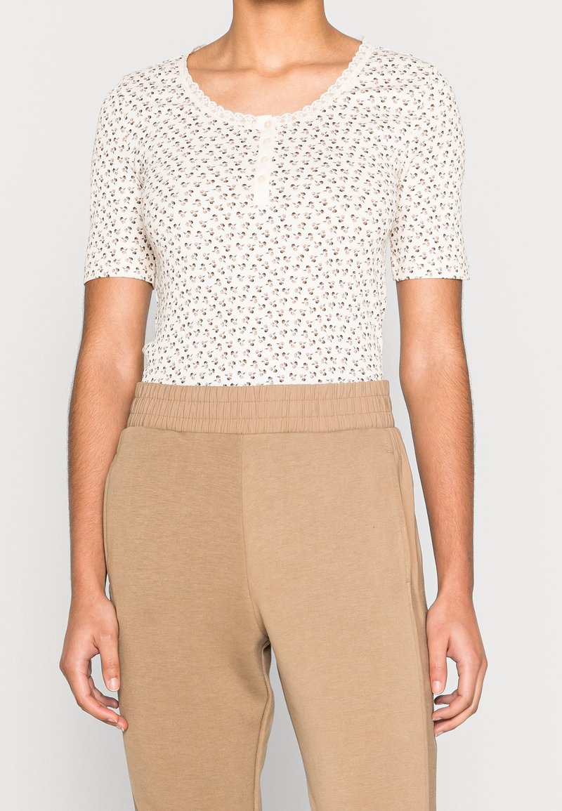Persona que lleva una camisa blanca de manga corta con estampado floral y botones, junto con pantalones beige de cintura alta y cintura elástica.