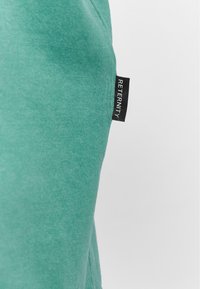 Grøn bomulds t-shirt med en glat tekstur og sideslit. Har en sort label med "RETERNITY" tekst på venstre side.