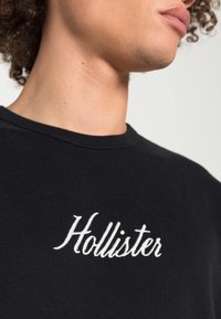 Černé bavlněné tričko s kulatým výstřihem, na levé horní části hrudi s bílým vyšívaným logem "Hollister". Hladký povrch.
