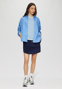 Hellblau gestreifte Button-Up-Bluse, blauer Pullover, marineblaue Cord-Rock, weiße Socken und silberne Sneakers. Das Modell steht mit den Händen in den Taschen.