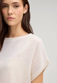 Leichter, cremefarbener Pullover mit lockerer Passform, U-Boot-Ausschnitt und kurzen Ärmeln. Weiche Textur und subtile Rippdetails an den Kanten.