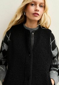 Svart sherpa-väst med en texturerad yta, med tryckknappsstängning och kontrasterande grå mönstrade tröjärmar. Rund halsringning.