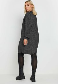 Abito in maglia a costine nera con maniche lunghe, collo alto e vestibilità ampia. Presenta un orlo arrotondato, indossato sopra collant neri e stivaletti.