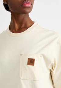 Lichtbeige t-shirt met een ronde halslijn, een zak met een bruine leren label en zilveren drukknoopdetails. Zachte katoenen textuur.