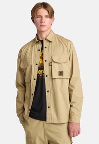 Beige button-up shirt gemaakt van stof met een gestructureerde afwerking. Beschikt over twee borstzakken, zwarte knopen en een logo-patch op de linkerzak.
