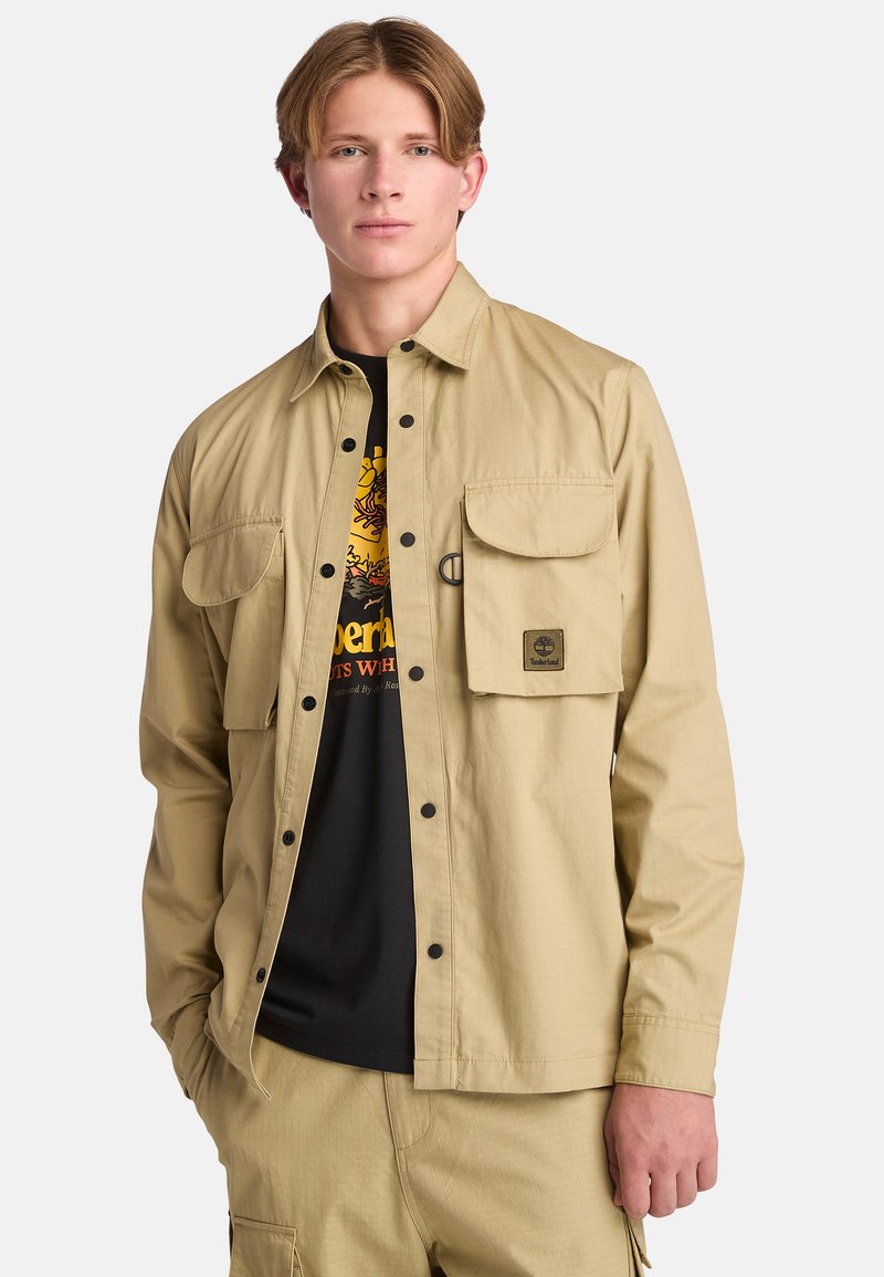 Beige button-up shirt gemaakt van stof met een gestructureerde afwerking. Beschikt over twee borstzakken, zwarte knopen en een logo-patch op de linkerzak.