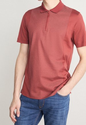 Mann trägt ein kurzärmeliges, strukturiertes dunkelrotes Polohemd mit halbem Reißverschluss, kombiniert mit blauer Jeans, steht mit einer Hand in der Tasche.