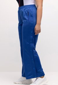 Pantalones deportivos azules con un bolsillo con cremallera, botones de presión en los lados, banda elástica en la cintura y una textura suave. Llevados con zapatillas deportivas blancas.