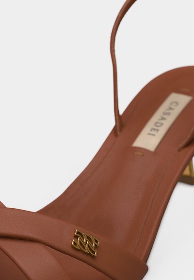 Καφέ δερμάτινο σανδάλι με λεπτά λουριά, ένα χρυσό διακοσμητικό έμβλημα στο μπροστινό λουρί και ένα μικρό τακούνι σε σχήμα τετράγωνου.