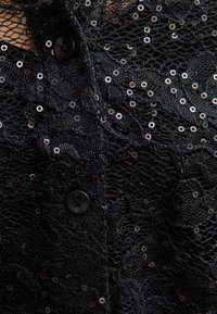 Czarna koronkowa tkanina z cekinami, z zapięciem na guziki i teksturowanym wzorem. Szczegółowy design z różnorodnym rozmieszczeniem cekinów.
