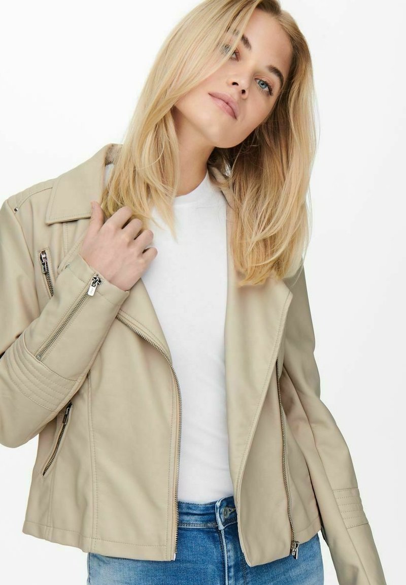 Beige imitert skinn bikerjakke med glidelåser, tettsittende design og rillede detaljer på skuldrene; brukt over en hvit t-skjorte og blå jeans.