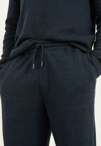 Nahaufnahme einer Person, die dunkelblaue gestrickte Sweatpants mit Zugband und Händen in den Taschen trägt.