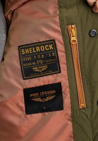 Grüne Jackenfutter mit einer pinken Innenschicht. Verfügt über Stoffetiketten: "SHELGROCK" in Gold und "PME LEGEND" in Schwarz mit goldenen Akzenten. Mit Reißverschluss versehenes Fach.