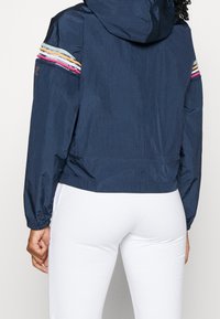 Námořnická mikina s kapucí a elastickými manžetami, zdobená vícebarevnými horizontálními pruhy na rukávech, vyrobená z lehkého, hladkého materiálu.