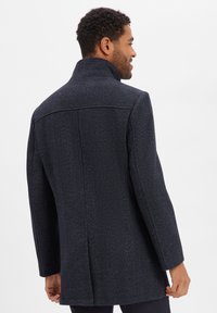 Dunkelblauer Mantel mit Fischgrätstruktur, mit hohem Kragen, langen Ärmeln und geradem Schnitt, von hinten betrachtet.