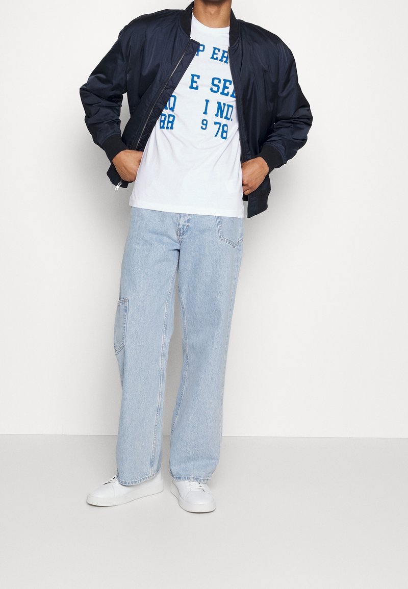 Navy bomber jacket with a zip front, white graphic t-shirt featuring blue text, light blue relaxed-fit jeans, and white sneakers.