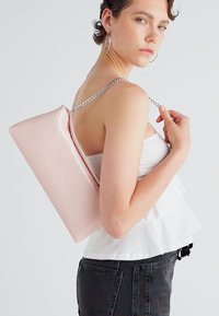 Rosa, glänzende, rechteckige Handtasche mit einer Kettenriemen, auf der Schulter getragen, ergänzt durch ein gerüschtes weißes Oberteil und dunkle Jeans.