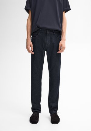 Homme portant un jean droit bleu marine, des chaussures noires et une chemise à manches courtes bleu marine, debout devant un fond uni.