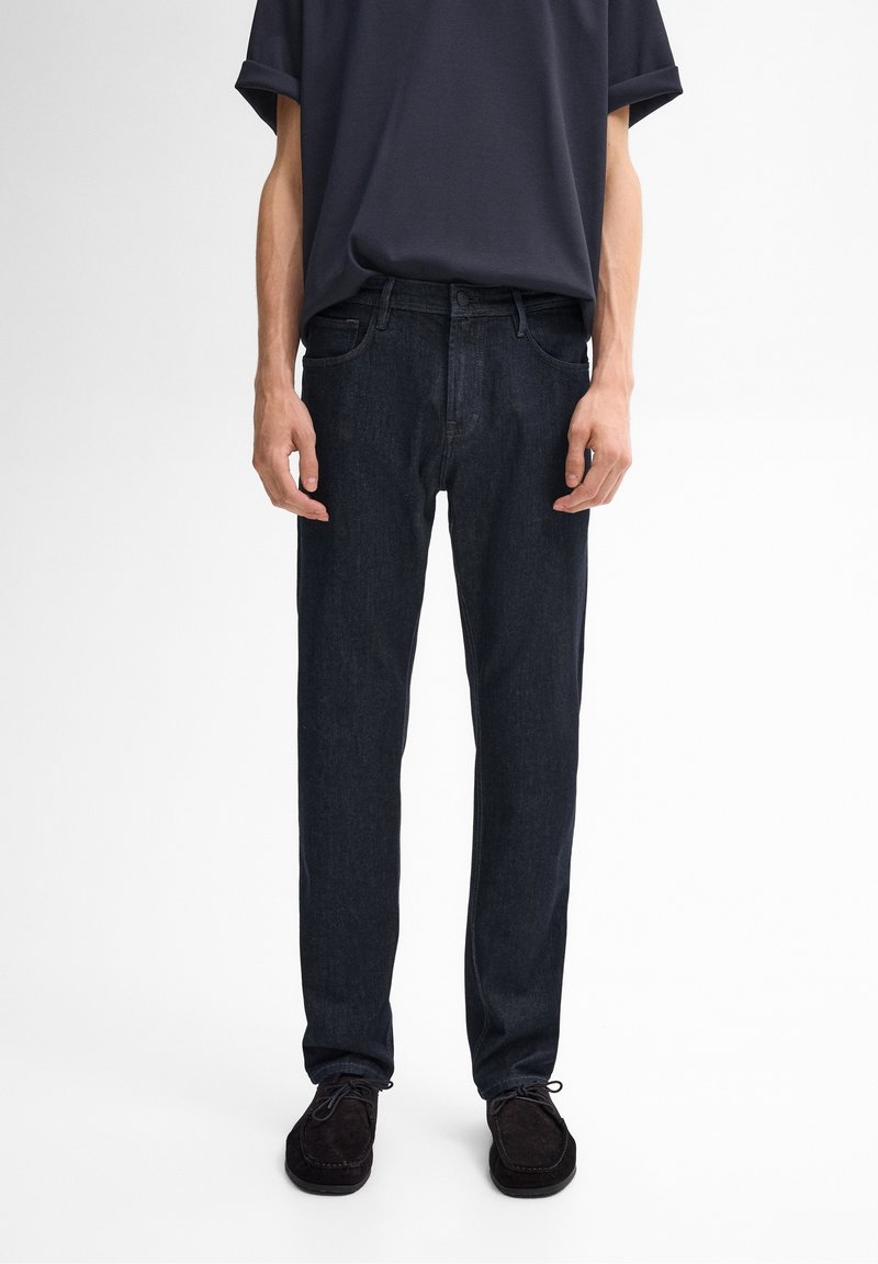 Homme portant un jean droit bleu marine, des chaussures noires et une chemise à manches courtes bleu marine, debout devant un fond uni.