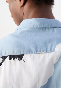 Világoskék ing, fekete-fehér absztrakt mintával a hátsó részén, gallérral és egy "Morato" felirattal ellátott fém brand címkével.