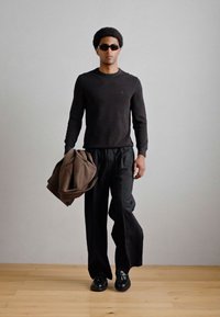 Černý svetr s dlouhým rukávem, volné černé kalhoty, černé boty a sluneční brýle, držící hnědý kabát. Hladká textura, ležérní design.