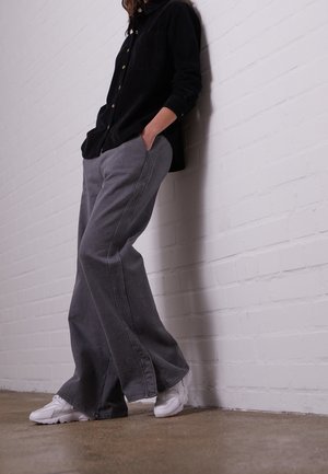 Personne appuyée contre un mur de briques blanches, portant une chemise noire à boutons, un pantalon large gris et des baskets blanches, les mains dans les poches.