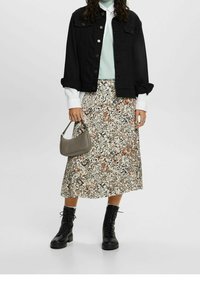 Personne portant une veste noire, un top bleu clair, une chemise blanche, une jupe midi fleurie, des bottes noires à lacets, et tenant un petit sac taupe.