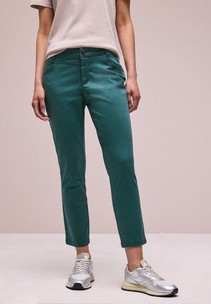 Osoba ubrana w turkusowe, dopasowane spodnie do kostek, beżową koszulkę i srebrne trampki, stojąca na jednolitej podłodze na tle neutralnego tła.