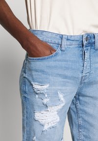 Jeans de mezclilla azul claro con áreas desgastadas y detalles deshilachados. Incluye una cinturilla, bolsillos frontales y un cierre de botón.