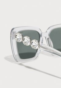 Lunettes de soleil carrées transparentes avec des verres foncés et trois grands cristaux clairs facettés sur la tempe. La monture est transparente avec des lignes épurées.