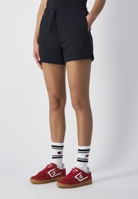 Zwarte katoenen shorts met zijzakken, een elastische tailleband met trekkoord, gecombineerd met rode suède sneakers en witte sokken met zwarte strepen.