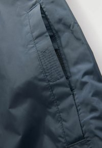 Veste imperméable bleu marine avec une texture lisse, dotée d'une poche latérale, de coutures visibles et d'un design élégant et épuré.