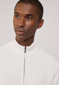 Maglione bianco a coste con zip, dotato di un colletto alto, caratterizzato da una zip nera e un tessuto testurizzato. Primo piano che mostra il collo e la parte superiore del corpo.