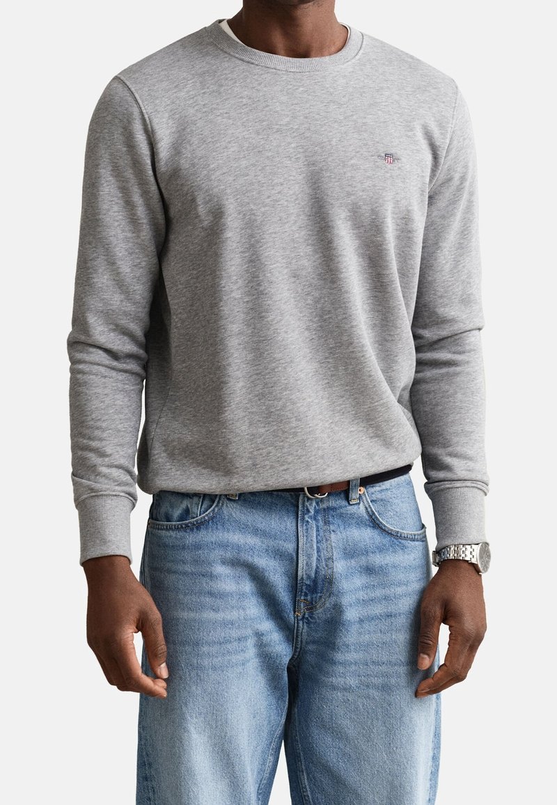 Homme portant un sweat-shirt gris clair à manches longues et un jean bleu clair, avec une montre argentée au poignet gauche.
