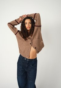 Braun gestrickte Strickjacke mit bordeauxfarbenen Streifen, ausgestattet mit Knöpfen und einer Fronttasche. Kombiniert mit dunkellblauen hoch-taillierten Jeans.