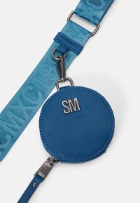 Blå cirkulær pung lavet af tekstureret stof, med et sølvfarvet "SM" logo. Fastgjort til et kontrasterende blåt bærehåndtag med et mønstret design.