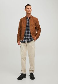 Giacca bomber in suede marrone, camicia a quadri bianca e nera, pantaloni cargo beige con tasche, scarpe nere con lacci, in piedi su uno sfondo neutro.