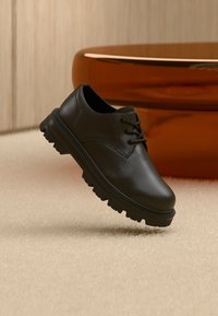 Sapato preto de couro com fecho de atacadores, bico arredondado e sola de borracha texturada em contraste. Fundo apresenta superfície texturada bege e objeto circular laranja.