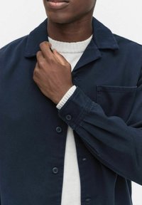 Marineblå skjorte med knapper og krave, udstyret med en brystlomme og manchetter, lagd over en struktureret cremesweater.