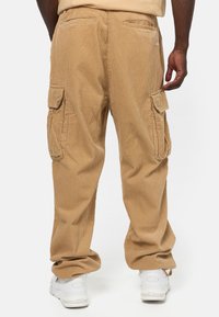 Vaaleanruskeat verryttelyhousut, joissa on sivutaskut, väljä istuvuus ja teksturoidunapintainen kangas. Vaatteet yhdistetty valkoisiin lenkkareihin, kuvattu takaa.