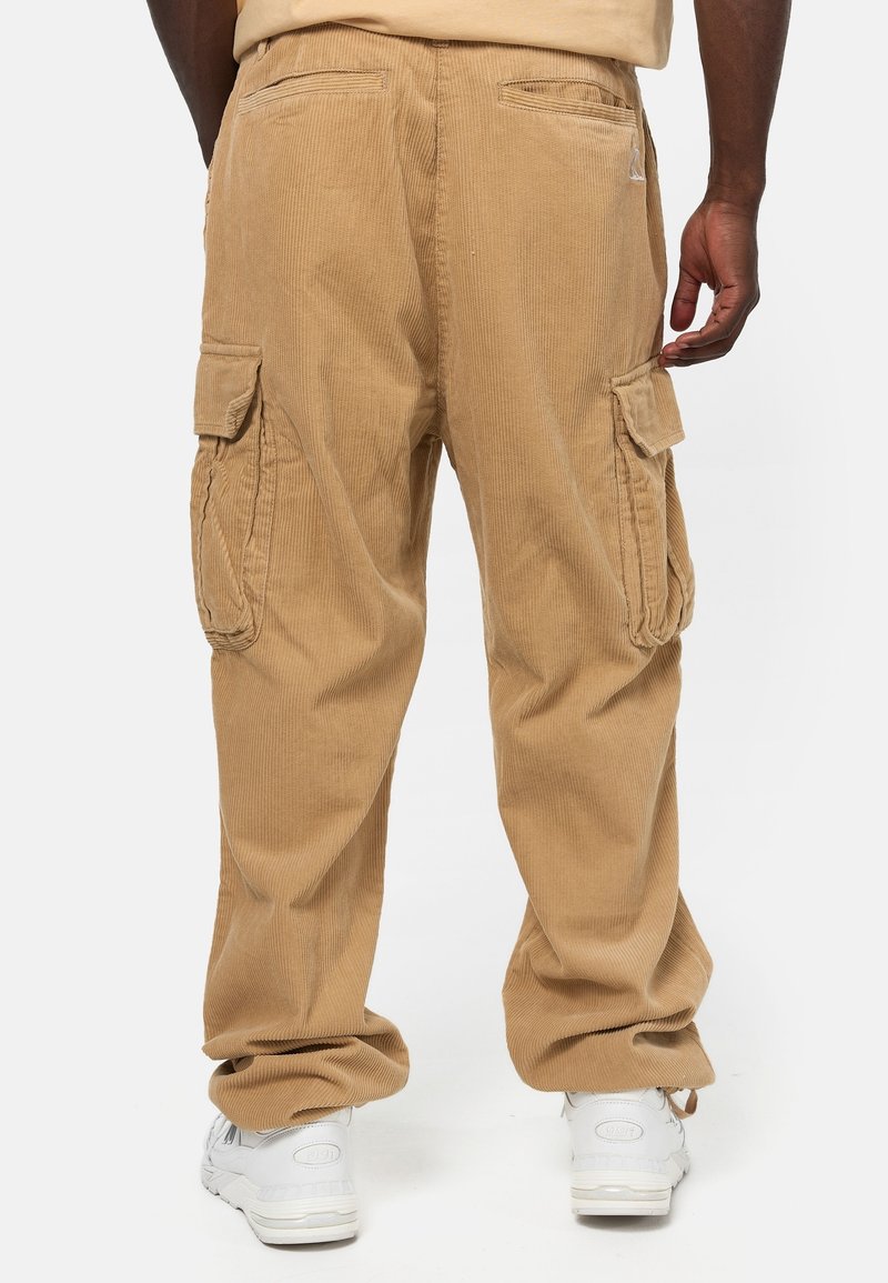 Vaaleanruskeat verryttelyhousut, joissa on sivutaskut, väljä istuvuus ja teksturoidunapintainen kangas. Vaatteet yhdistetty valkoisiin lenkkareihin, kuvattu takaa.