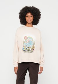 Gaiši rozā džemperis ar apaļu izgriezumu un nolaistiem pleciem. Uz tā ir krāsains grafisks dizains ar sauli, viļņiem un ziediem.