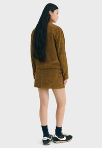 Frau mit langen schwarzen Haaren, die eine braune Cordjacke und einen Rock trägt, schwarze Socken und schwarz-weiße Nike-Sneaker, die seitlich steht.