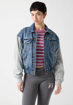 Młoda kobieta z krótkimi ciemnymi włosami, ubrana w dżinsową kurtkę z szarymi rękawami, paskowany fioletowy krótki top i szare legginsy z białym grafiką skrzydeł.