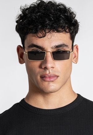 Mladý muž s kučeravými tmavými vlasmi, s malými obdĺžnikovými čiernymi slnečnými okuliarmi so zlatými rámami a čiernym rebrovaným tričkom na jednoduchom pozadí.