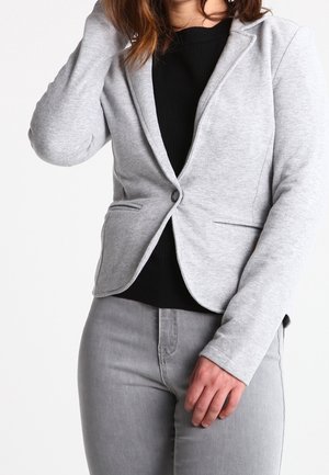 Žena v světle šedém blejzru přes černý top a světle šedých džínách, s jednou rukou u obličeje a druhou podél těla.