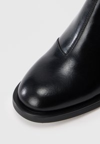 Bout de chaussure en cuir noir brillant avec une couture détaillée et un bord de semelle lisse et arrondi sur un fond clair.