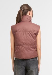 Vesta bez rukávov v tlmenej hnedej farbe so zvýšeným golierom a horizontálnym prešívaním; doplnená tmavosivými nohavicami.
